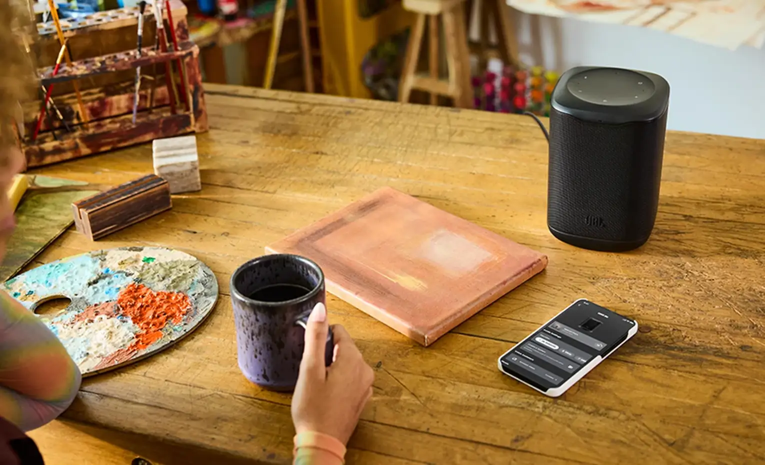 اسپیکر جی بی ال Cove M1؛ بررسی امکانات و کیفیت صدا اسپیکر JBL Cove M1 کنار گوشی، کتاب و لیوان روی میز